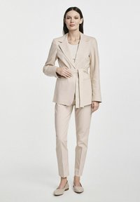 Beżowa marynarka dopasowana z wiązaniem w talii, w zestawie z wąskimi spodniami i pasującymi beżowymi balerinami, charakteryzująca się gładką fakturą i minimalistycznym stylem.