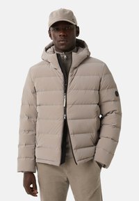 Beige puffer jacket with horizontal stitching, hood, and black zipper details. Worn over a grey sweater, paired with light-colored pants.