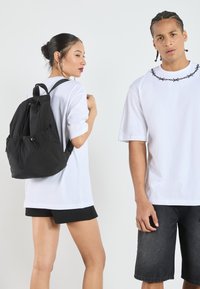 Svart dragsäck med justerbara remmar, tillsammans med en löst sittande vit t-shirt och svarta shorts. Slät tyg utan märkbara mönster.