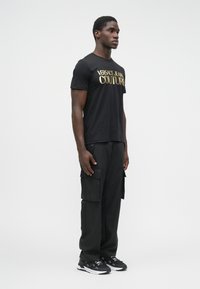 Vīrietis stāv, ģērbies melnā Versace Jeans Couture t-kreklā, melnos kabatbiksēs un melnbaltās sporta kurpēs, pret vienkāršu baltu fonu.