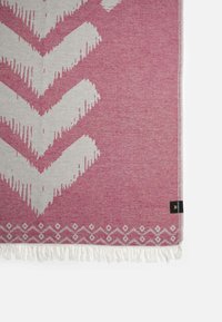Manta tejida en tonos rosa y crema con un patrón de chevron y flecos blancos. Presenta un diseño de borde diagonal en forma de diamante. Etiqueta en el lateral.