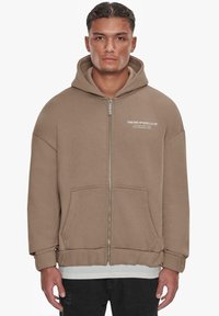 Brown zip-up hoodie with a large hood, front pockets, and ribbed cuffs. Features white text on the left chest area. Soft, textured fabric.