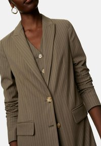 Olivově zelené sako s proužky, dlouhými rukávy, klopovým límcem a dvěma předními kapsami; na přední straně mají světlé knoflíky.