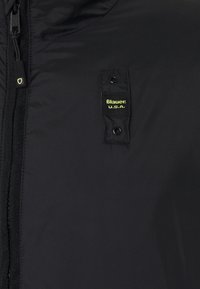 Veste noire isolante avec fermeture à glissière et boutons-pression. Présente une étiquette jaune cousue portant l'inscription "Blauer : U.S.A." sur la poitrine.