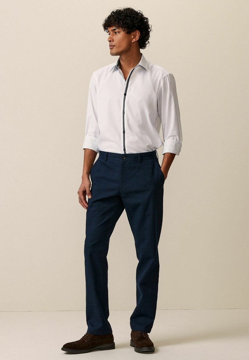 Weißes Hemd mit Button-Down-Kragen und gemusterten Manschetten, kombiniert mit navyblauen Hosen und braunen Schuhen. Schmaler Schnitt mit modernem Design.