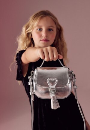 Mladá dívka se světlými vlasy drží malou stříbrnou kabelku se srdcovým zapínáním a třásní na růžovém pozadí.