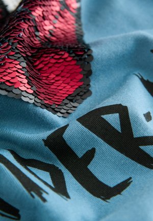 Tissu bleu avec un motif texturé de sequins et des écailles noires et rouges. De grandes lettres noires partiellement visibles à la surface.
