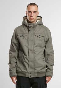 Chaqueta verde oliva con capucha, botones frontales, dos bolsillos en el pecho, bolsillos laterales y puños y dobladillo acanalados. Forro interior suave visible.