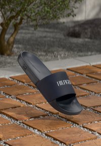 Schwarze Tommy Hilfiger Badesandalen, die über einem Holzblock und einem Kiesboden im Freien schwebt, mit einem grünen Baum im Hintergrund.