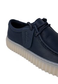 Scarpa in pelle navy con punta tonda, superficie testurizzata, lacci e suola traslucida con scanalature. Presenta un'etichetta blu con il marchio "Clarks".