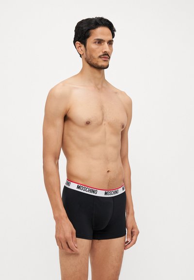 Sorte boksershorts med en hvit og rød Moschino-logo på kanten. Glatt tekstur, tett design, lengde til midten av låret. Modell vist fra siden.