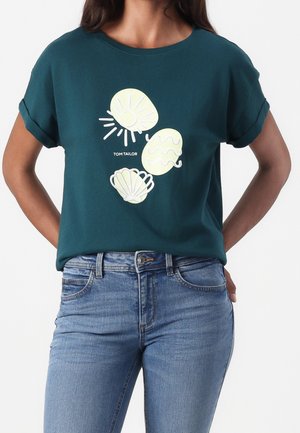 Femme portant un t-shirt bleu sarcelle à manches retroussées avec des motifs abstraits jaunes représentant une coquille et le soleil, associé à un jean bleu clair.