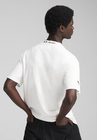 Hvid bomulds t-shirt med rund hals, korte ærmer, og et sort logo på halsen og ærmet. Enkel, afslappet pasform.