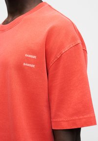 T-shirt en coton corail à manches courtes, arborant deux logos blancs "SAMSSØE" sur le côté gauche, avec un col rond et une coupe décontractée.