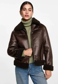 Jachetă din piele maro cu guler și manșete din blană, închidere cu fermoar și design scurt. Purtați-o peste un pulover verde cu guler înalt ribbat.