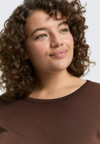 Bruin, kort mouwtop met een ronde halslijn. Zachte, gladde textuur. Het model heeft krullend haar en toont alleen het gezicht en het bovenste deel van de torso.