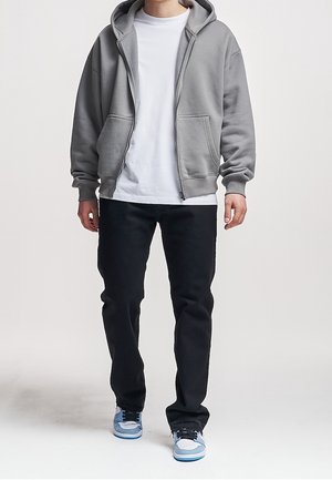 Person iført en grå lynlåshoodie, hvid t-shirt, sorte jeans og blå-hvide sneakers, stående mod en ensfarvet baggrund.