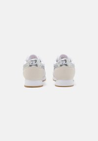 Zapatillas blancas con acentos plateados, textura de tela suave, cuello acolchado, logo en el talón y una suela de goma beige.