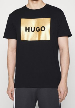 Homme portant un t-shirt noir à manches courtes avec un patch rectangulaire doré brillant et un texte noir "HUGO" sur la poitrine.
