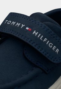 Σκούρο μπλε ναυτικό, μοκασίνι σουέτ με φαρδιά λουρί με βέλκρο. Διαθέτει άσπρα ραφές και το λογότυπο "TOMMY HILFIGER" με τριχρωμία.