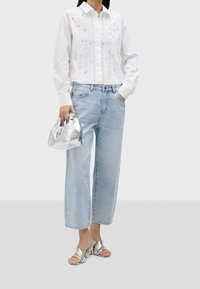 Witte button-up shirt met plooien en versieringen, lichtblauwe wijde jeans, zilveren metallic tas en strappy zilveren sandalen.