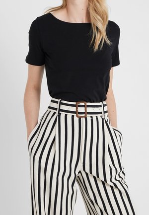 Femme portant un haut noir à manches courtes rentré dans un pantalon blanc taille haute à rayures verticales noires et une ceinture assortie avec une boucle carrée.
