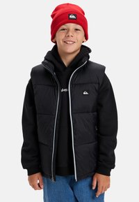 Skrattande pojke iklädd en röd mössa, svart vadderad väst, svart hoodie och blå jeans som står framför en neutral bakgrund.