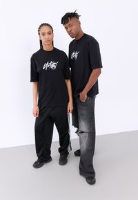 Svarta t-shirts med en vit grafisk logo bärs tillsammans med lösa svarta träningsoverallsbyxor med vita ränder och slitna grå jeans.