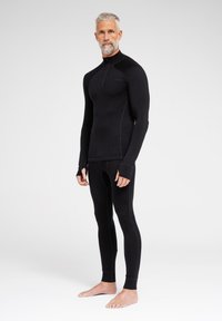 Schwarzes thermisches Basisschicht-Outfit, bestehend aus einem langärmeligen Oberteil mit einem halben Reißverschluss und Daumenlöchern sowie enganliegenden Leggings mit glatter Textur.