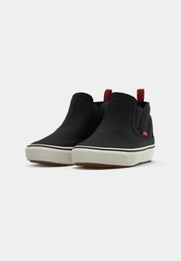 Black slip-on shoes with a smooth texture, elastic side panels, white rubber sole, and red checkered pull tab.