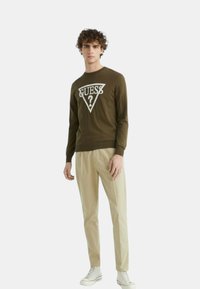 Maglione verde a maniche lunghe con un logo triangolare bianco con la scritta "GUESS?", abbinato a pantaloni beige e sneakers bianche.