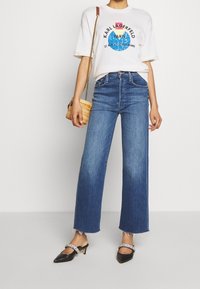 Bílý grafický tričko s modrým a růžovým designem, vysoké modré džíny s širokými nohavicemi, černé boty s špičatou špičkou a malá pletená taška.