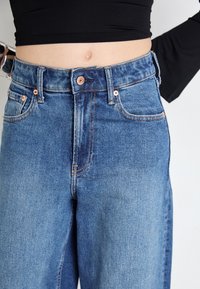 High-waisted blue denim jeans with copper-tone hardware, five-pocket design, and contrasting stitching. Smooth texture, straight-leg cut.