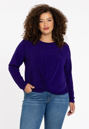 Lilla langærmet top med en twistknude foran, lavet af blødt stof, parret med lyseblå jeans.