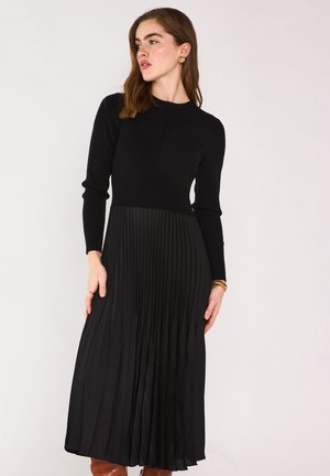 Femme aux cheveux bruns ondulés portant une robe noire plissée à manches longues, des boucles d'oreilles créoles dorées et des bottes marron montant jusqu'aux genoux, regardant vers la gauche.