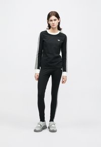 Mladá žena stojí oblečená v čiernom Adidas teplákovom súprave s bielymi pruhmi a šedých športových topánkach na jednofarebnom bielom pozadí.