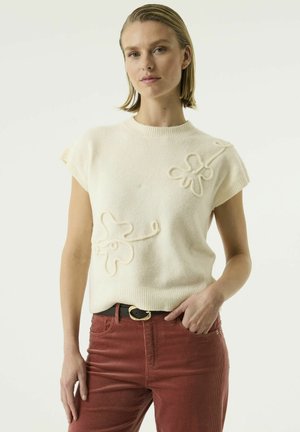 Femme portant un pull à manches courtes crème avec des motifs floraux texturés et un pantalon en velours côtelé couleur rouille avec une ceinture noire.