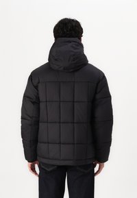 Zwarte pufferjack met een capuchon, voorzien van een geb quilt ontwerp en een gestructureerde afwerking. De mouwen zijn iets oversized, wat zorgt voor een ontspannen pasvorm.