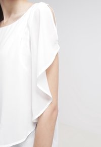 Blouse blanche avec une coupe décontractée, en chiffon doux et à manches volantées. Le design comprend un col rond et une texture lisse.