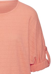 Hellkorallenfarbener Pullover, aus strukturiertem Stoff gefertigt. Mit Rundhalsausschnitt und umgeschlagenen Ärmeln mit Knopf-Details.