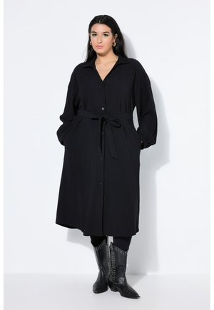 Kobieta z długimi ciemnymi włosami, ubrana w czarną sukienkę midi z paskiem i guzikami oraz czarne, ćwiekowane kozaki do połowy łydki, stojąca na białym tle.