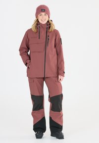 Skijacke und Hosen-Set in gedämpftem Rosa und Schwarz, aus synthetischem Material. Die Jacke hat eine Vorderentasche; die Hose verfügt über Utility-Taschen und Farbblockierungen.