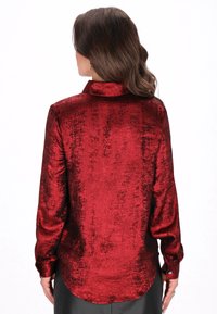 Rød metallisk skjorte med lange ærmer, krave, glat tekstur og manchetter med knapper. Har en rundet kant og en skinnende overflade.