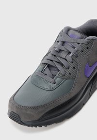 Γκρι αθλητικά παπούτσια Nike με μωβ σήμα, υφασμάτινες επιφάνειες από δέρμα με υφή και σουέτ, γκρι κορδόνια και επενδεδυμένο κολάρο σε λευκό φόντο.