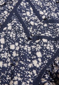 Námořnická tkanina s bílým květinovým vzorem, obsahující knoflíky a dekorativní krajkový lem podél švů, což vytváří texturovaný vzhled.