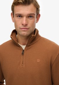 Brauner Zip-Pullover aus geripptem Material mit hohem Kragen, ausgestattet mit einem kleinen Logo auf der Brust und einer hellen inneren Schicht.