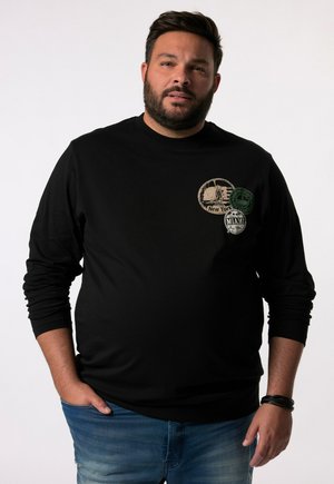 Sudadera negra de manga larga, con parches circulares en crema y verde, que muestran nombres de ciudades. Ajuste estándar con puños acanalados.