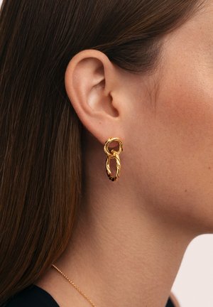 Pendiente de aro de doble eslabón retorcido en oro, llevado en la oreja izquierda de una mujer con cabello castaño lacio visible.