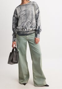 Sudadera oversized en gris y negro con diseño abstracto, combinada con jeans de pierna ancha de color verde claro y un pequeño bolso negro.