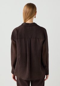 Camicia marrone, comoda e realizzata in tessuto leggero. Presenta un colletto, maniche lunghe e polsini con bottone, con una texture semplice e un dettaglio di yoke sul retro.